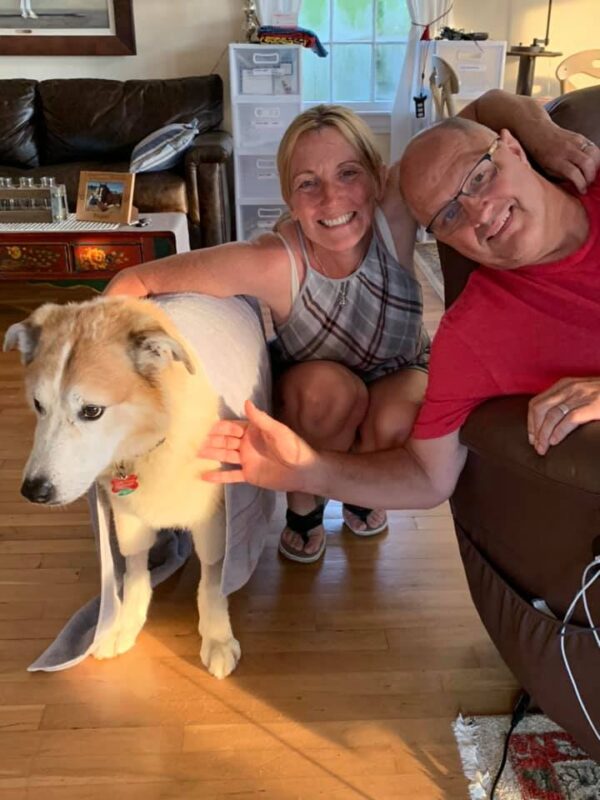 A man, woman, and dog inside the living room of a house. The man and woman are both smiling and have their hands on the dog. The man is leaning into the photo from the right side of the frame, while the woman is in the center, squatting down to be at the dog's level. The dog looks off to the left side of the frame.