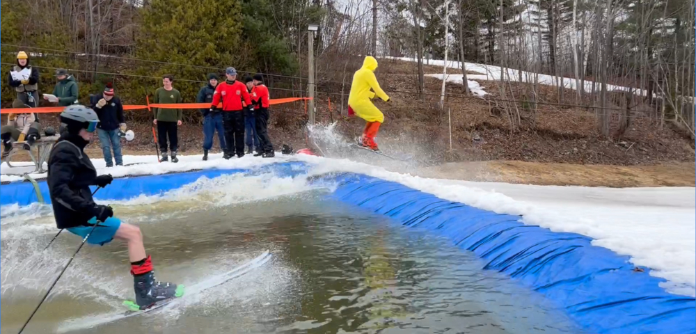 Two people on skis attempt to cross a slush pit lined with a blue tarp. Several spectators are visible in the background.